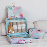 A pastel floral travel toiletry organizer set displayed on a white textured blanket, featuring a hanger pouch, an open zippered organizer with shampoo and body wash bottles, and a smaller matching pouch.