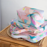 Two pastel floral toiletry and cosmetic bags stacked on a woven rattan tray, with a light pink background and a small pink Mindful Marlo brand patch on the top pouch.