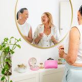 Two women laughing in front of a mirror with a pink jewelry box on a table.